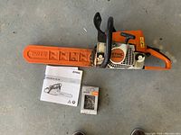 Top-down view of chainsaw with orange bar cover, operator's manual, and genuine parts box
