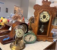Four mantle clocks displayed together on a kitchen countertop with two original winding keys visible; includes an ornate carved wooden Ansonia gingerbread clock, a brass anniversary clock under glass dome, a small green oval clock, and a wooden-cased clock lying flat.