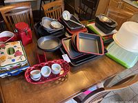 Wide view of table-top with stacked baking trays, pans, ceramic dishes, red canister, plastic cover, and other kitchen items.