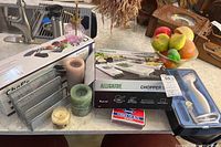 Photo of items on marble kitchen counter including boxed mandoline slicer, Alligator chopper box, candles, metal organizer, and glass bowl with faux fruit.