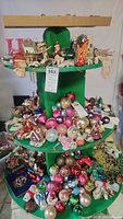 Three-tier green ornament stand filled with assorted Christmas baubles, feathers, and various holiday figures.