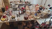 Wide view of tabletop filled with various decorative items including figurines, dolls, glassware, and holiday decor.