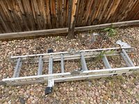 Photo of aluminum ladders on gravel next to a wooden fence with some weeds around.