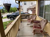 View of two light-colored wicker rocking chairs with red cushions on a porch along with a round dark red wicker table and hanging wicker planters.