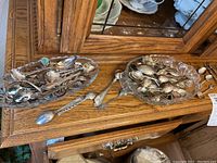Two glass dishes and loose spoons spread out on a wooden cabinet surface showing detailed souvenir spoons with mixed metals and decorative handles.