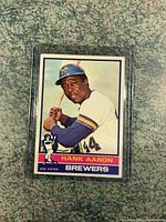 Front side of 1976 Hank Aaron baseball card showing Hank Aaron in Milwaukee Brewers uniform holding a bat, card encased in plastic sleeve.