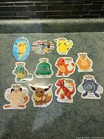 Eleven 1999 Pokémon stickers arranged on a green surface showing all characters including Pikachu, Ash, Brock, and original starter Pokémon.