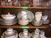 Photo showing the full set arranged on a shelf including stacked plates, bowls, multiple cups and saucers, and serving ware. Pattern is small pink roses with green foliage and gold trim
