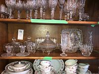 Shelf with various crystal and glass serving pieces including covered dishes, plates, bowls, and glasses.
