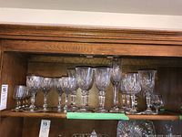 Clear crystal stemware glasses with cut and etched designs displayed on a wooden shelf, showing various sizes and shapes.