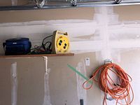 Electrical cords, booster cables, and blue insulated lunch bag on garage wall shelf and hooks.