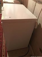 Photo showing side view of white Frigidaire chest freezer in basement setting with visible power cord and part of carpeted floor.