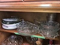 Shelf containing various glass serving bowls and plates, some stacked, showing clear glass with patterns and white porcelain serving ware.