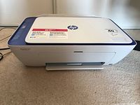 Front view of HP Deskjet 2655 all-in-one printer showing white and blue plastic casing on carpeted floor.