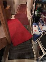 Photo showing several mats and runners arranged on basement floor with shelving and household supplies in background. Red mat prominently visible with rounded corner and stain.