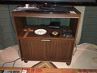 Front view of wooden media stand with various items on open shelf and top surface, showing vertical panel sliding doors and visible caster wheels.