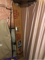Tall unopened cardboard box with visible water damage at bottom, containing mirror panels, positioned in basement next to pipes and curtain.