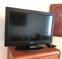 Front view of RCA television on a wooden cabinet with remote and a small box to the right.