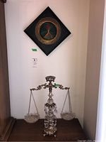 Decorative brass scale with two hanging glass dishes placed on a wood surface below a framed wire string art piece hanging on a wall.