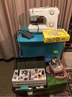 Full lot view showing Singer Prelude sewing machine on blue storage bin, sewing supply case open with multiple compartments holding thread, needles, bobbins, scissors, and small tools, sewing book titled 'The Family Sewing Book' next to machine.