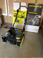 Side view of Yardworks electric snow blower with packaging box seen behind it in garage setting.