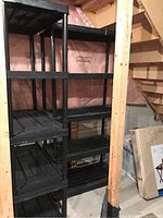Two black PVC utility shelving units, five-tiered, shown standing side by side in basement space against a wall with pink insulation visible. Shelves and tubular legs visible with vented slats on each shelf.