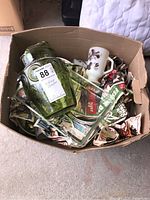 View of vintage glassware including green vase, clear baking dish, and cartoon character mug arranged in cardboard box packed with newspaper.