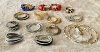 Fourteen pairs of hoop style pierced earrings in various sizes arranged on a light cloth background. Styles include textured gold-tone hoops, smooth gold hoops, silver-tone hoops with beads, colorful beaded hoops in blue and red, and black and silver accented hoops.