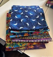 Stack of 10 pieces of folded quilting fabric with one piece showing blue fabric with moon and stars pattern