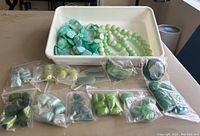 Photo showing a white tray with loose beads strings and multiple small plastic bags with various green and blue beads of different shapes and sizes.