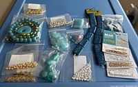 Photo showing assortment of beads and jewelry findings in multiple plastic bags, including turquoise, gold-tone, and silver beads, and two fabric bead holder straps.