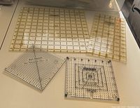 Photo showing six transparent cutting rulers on white surface. Two square rulers in front and four rectangular rulers at back. The rulers have measurement grids and labels.