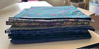 Stack of 10 folded quilting fabric pieces showing various patterns and colors dominated by blues and greens.
