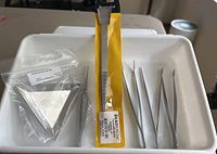 Jewelry making tools on a white tray including eight metal triangular trays, four pairs of tweezers, a bead shovel, and one metal rod tool.