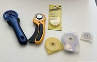 Photo of two rotary cutters (blue and orange/black) with four additional rotary cutter blades in various transparent cases and one unopened pack of OLFA blades.