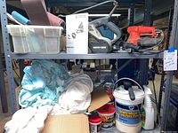 Wide view of shelving showing two electric sanders, plastic containers with sanding belts, instruction manual, wall repair and painting supplies like concrete patch and spray bottles.