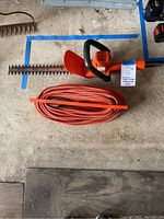 Top view of Black & Decker hedge trimmer and orange extension cord laid on a concrete floor.