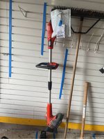 Full length view of Black & Decker cordless weed whacker along with garden tools and broom in background.