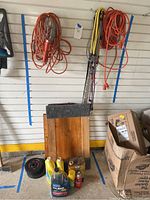 Garage wall with bundled orange extension cords including one with a hanging cage work light, assorted colorful bungee cords, a dolly with carpeted surface, wooden cutting board, black weights, and assorted cleaning liquids on the floor.