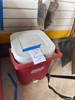 Full view of the red Coleman beverage cooler with white lid and attached carrying handle, placed under a garage shelf with gardening tools in background.