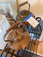 View of two vintage copper teapots on a metal rack, showing one teapot with round body and the other with wider squat body, both featuring wooden handles and lids with wooden knobs.