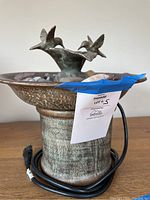 Photo showing the full fountain with electrical cord coiled at the base, featuring a pedestal style base with wide rim and two hummingbirds perched on flower-shaped basin.
