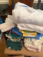 Stack of folded linens including flannel bed linens, kitchen towels, pot holders, tablecloths, and napkins in various colors and patterns.