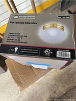 Box of Boston Harbor single flush mount ceiling fixture with polished brass finish and frosted glass shade visible on top of larger box.