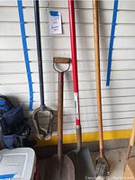 Post hole digger, shovels, and edger standing upright against wall with garage background