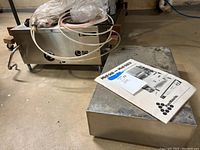 Stainless steel distiller unit with attached tubing, plastic faucet/spout, and user manual on concrete floor.