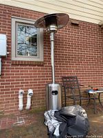 Full view of patio propane heater standing near brick house wall. The heater has a bent top canopy and is accompanied by a folded zippered storage cover on the ground.