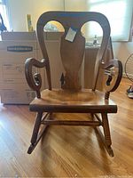 Front-facing view showing maple-tone wooden rocking chair with damaged veneer on seat and backrest, scroll-shaped armrests, and overall wear.