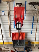 Large red snow shovel with wide blade and black handle grips alongside other shovels hanging on garage wall