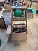 Front view of wooden step ladder with two slatted steps with visible wear and paint splatters, showing box of assorted hand tools underneath.
